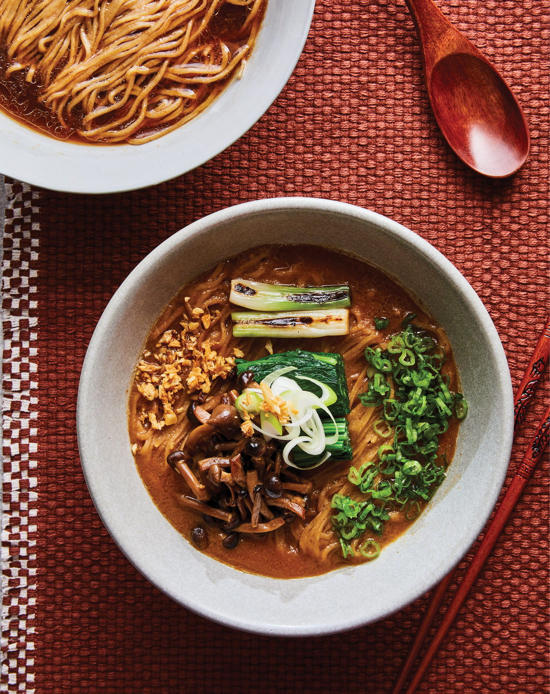 Bowl of ramen with noodles, vegetables, and a wooden spoon on a textured red surface.