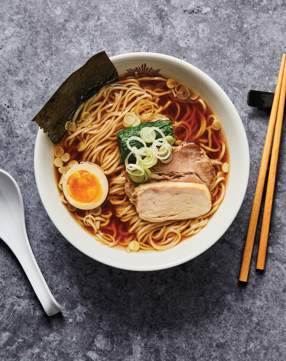 Bowl of ramen with noodles, egg, and pork on a gray surface with chopsticks and a spoon.