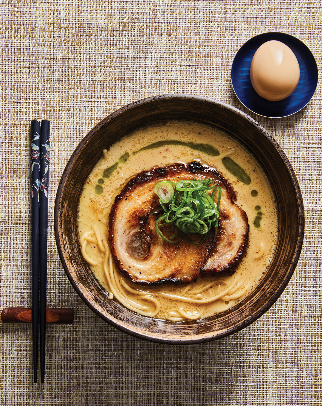 Bowl of ramen with pork slices and green onions on a textured surface with chopsticks and a ramen egg.