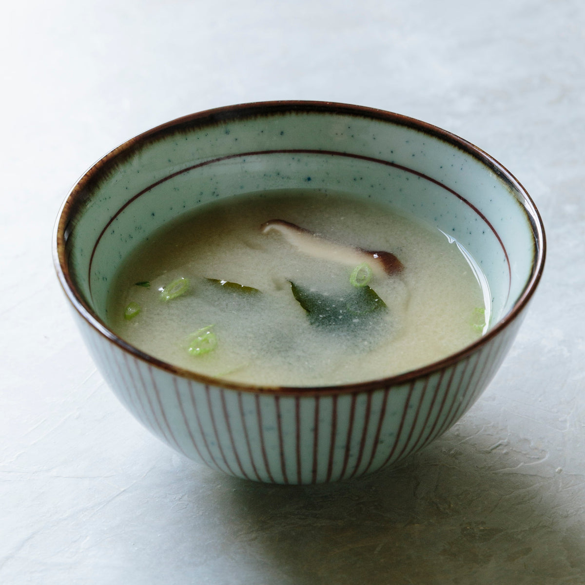 A ceramic bowl filled with miso soup, featuring a pale green interior, brown speckled rim, and vertical brown stripes on the exterior, set on a light gray textured surface.