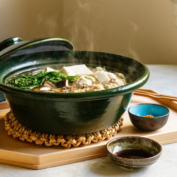 Three quarters view of green Japanese donabe with steaming chicken mizutaki hotpot in it. Next to that a small bowl with red yuzu kosho, a bowl of soy sauce and serving chopsticks.