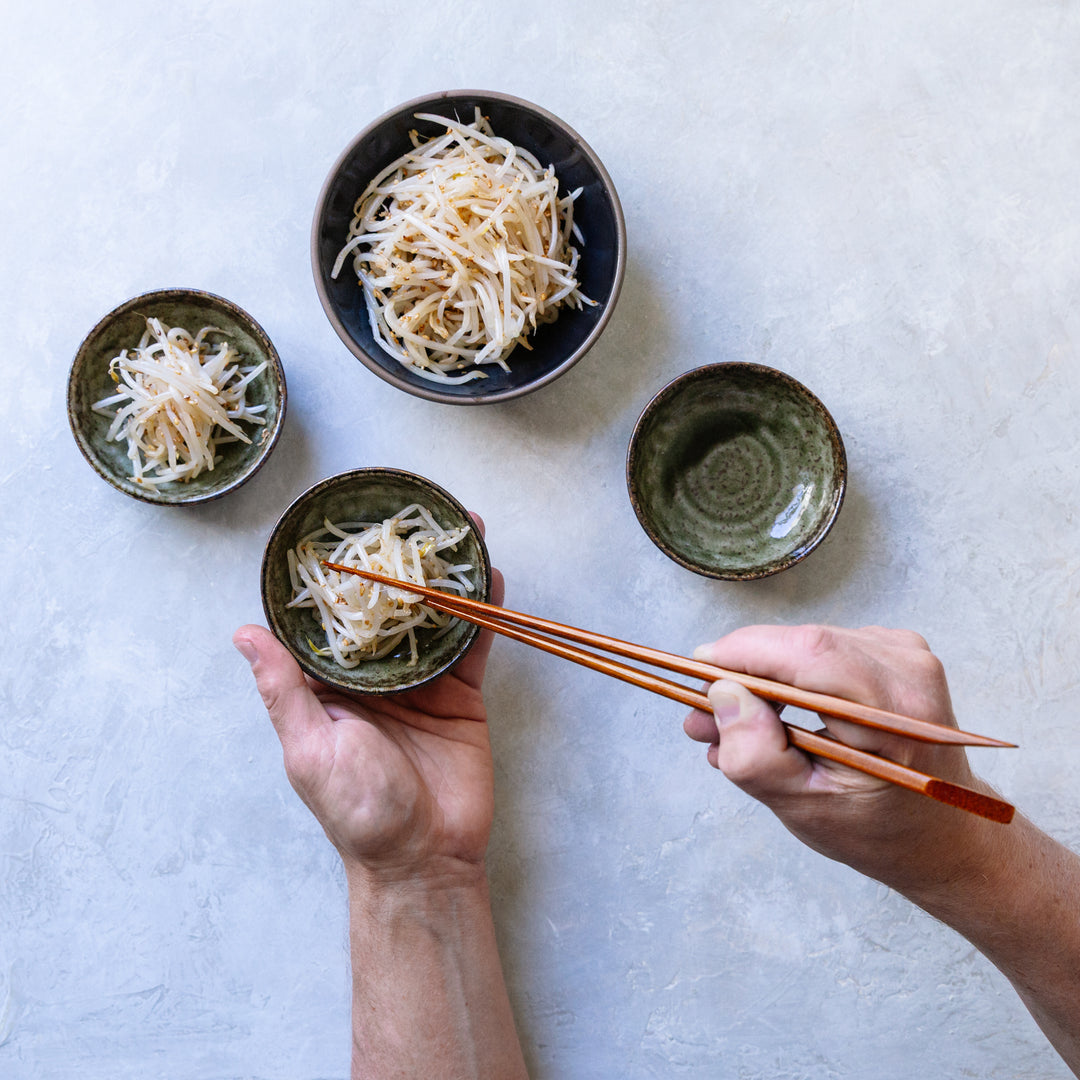 Four small bowls with noodles on a light gray surface, with hands using chopsticks.