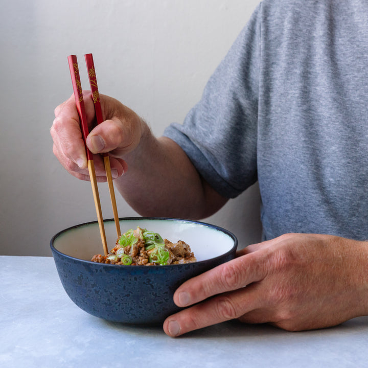 Person holding a blue bowl with food using chopsticks on a light surface.