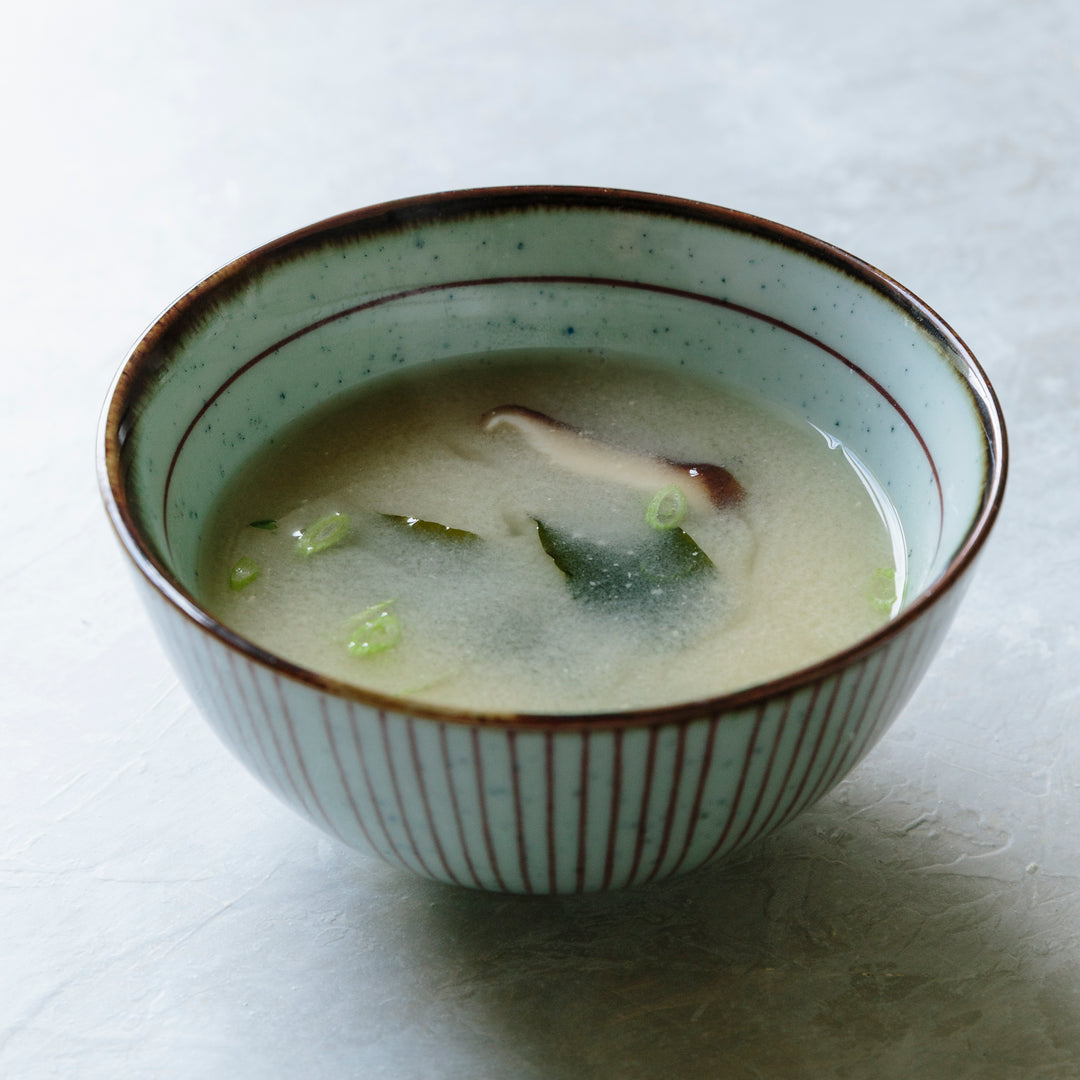A ceramic bowl filled with miso soup, featuring a pale green interior, brown speckled rim, and vertical brown stripes on the exterior, set on a light gray textured surface.