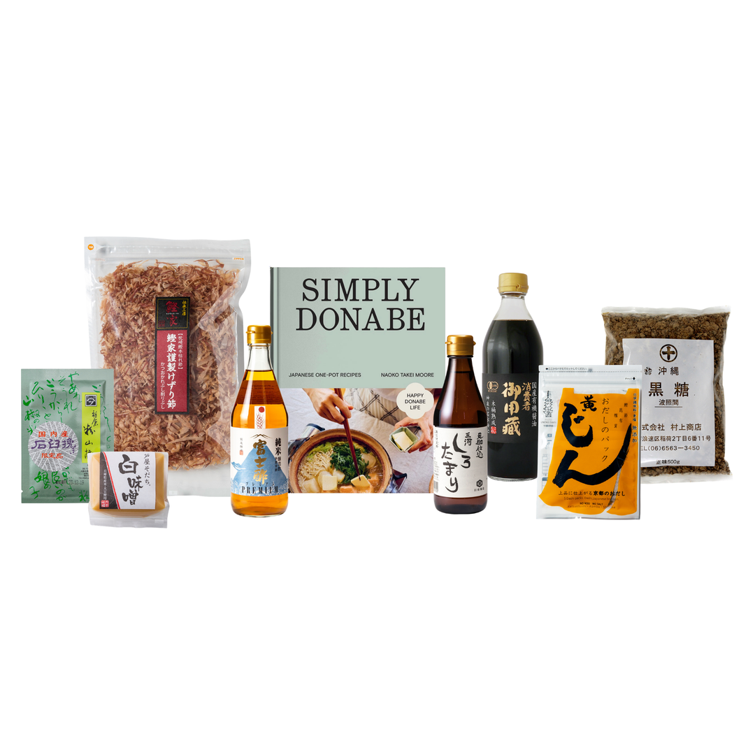 A curated assortment of Japanese food products arranged on a white background, including packaged rice, soy sauce bottles, a cookbook titled 'SIMPLY DONABE,' and other grocery items.