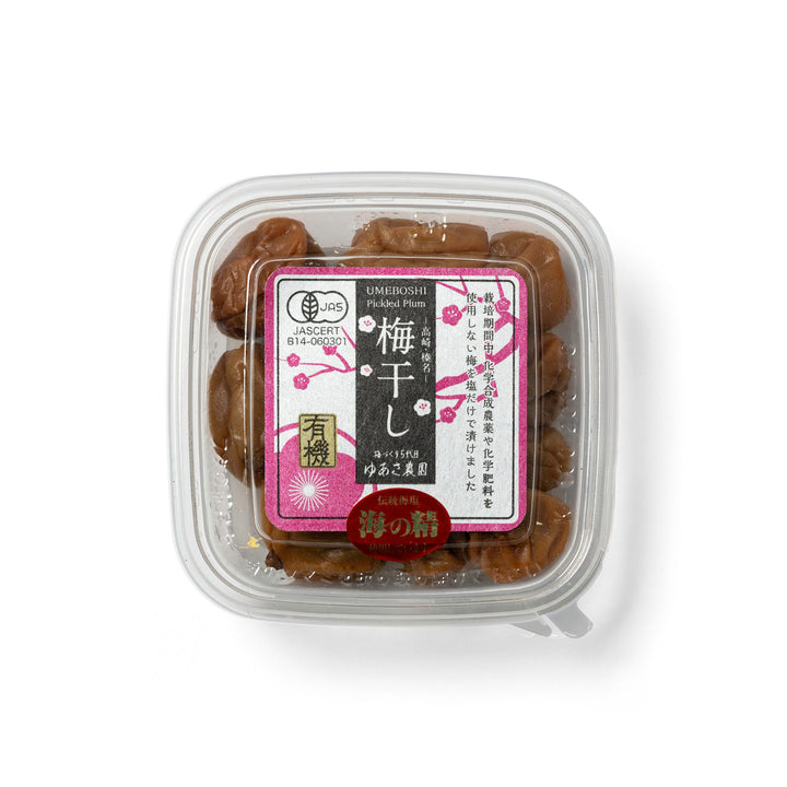 A clear plastic square container holding several dark brown plum dates, with a decorative Japanese label on top. 