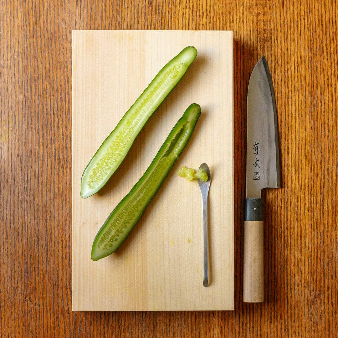 Large wooden cutting board with two halves of a sliced cucumber and a spoon that has scooped out some of the cucumber seeds with a Japanese chefs knife next to the cutting board.