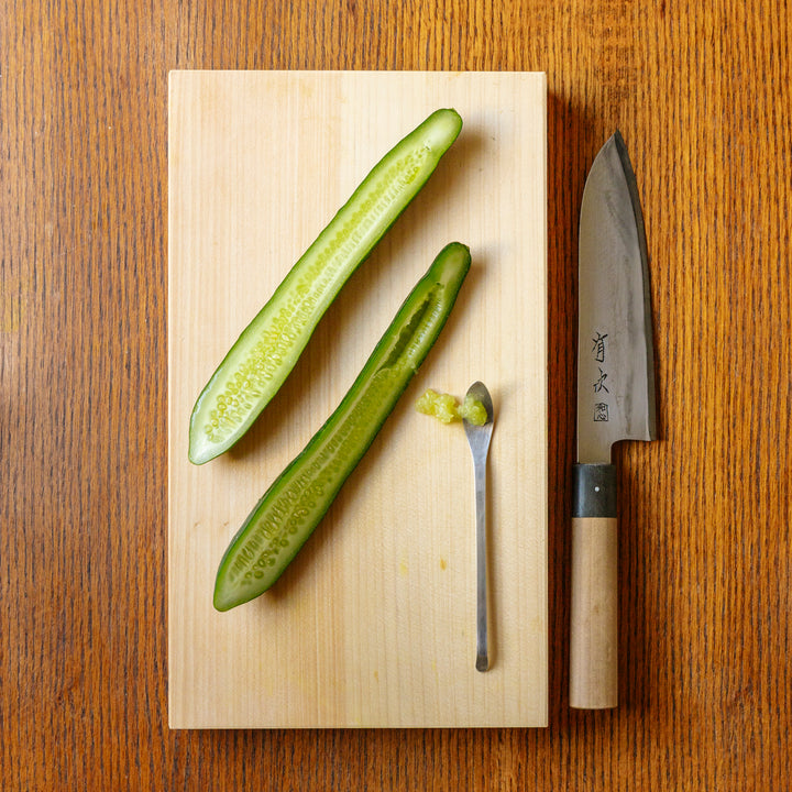 Large wooden cutting board with two halves of a sliced cucumber and a spoon that has scooped out some of the cucumber seeds with a Japanese chefs knife next to the cutting board.